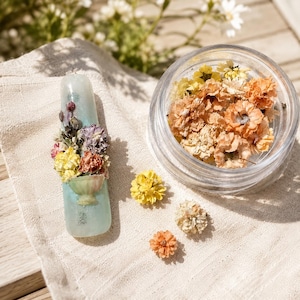 Könnte beinhalten: Eine hellblaue Kerze mit einem Blumengesteck in einer kleinen Vase. Die Anordnung umfasst gelbe, violette und orangefarbene Trockenblumen. Ein klares Glas mit Trockenblumen und mehrere lose Blüten sind ebenfalls vorhanden.