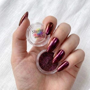 May include: A hand holding two small, clear jars of nail art supplies. One jar contains a dark, shimmery powder, and the other has the text "Starry nails" on the lid. The nails are painted a metallic burgundy color.