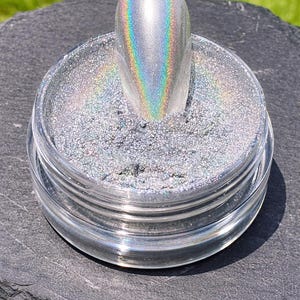 Könnte beinhalten: Ein klares Glas mit silbernem Glitzer und einem holografischen Nagel. Der Nagel hat einen Regenbogen-Schimmer, der verschiedene Farben reflektiert. Das Glas steht auf einer dunkelgrauen, strukturierten Oberfläche, mit grünem Gras im Hintergrund.