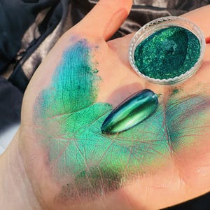 May include: A hand displays green and blue iridescent nail polish and loose glitter. A clear container holds the glitter, and a single, polished nail is also shown. The palm is dusted with the same shimmering pigments, creating a vibrant, eye-catching display.