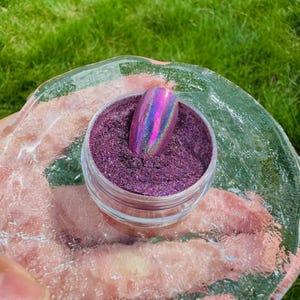 May include: A small, clear jar filled with shimmering purple powder, with a holographic nail art sample. The jar sits on a clear, textured surface, with green grass in the background. The nail art sample has a metallic, iridescent finish.