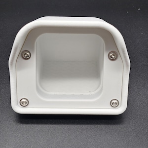 Puede incluir: Un componente de plástico blanco rectangular con bordes redondeados y un interior empotrado. Cuatro pequeños pernos plateados aseguran el componente. El objeto parece ser una carcasa o un soporte de montaje.