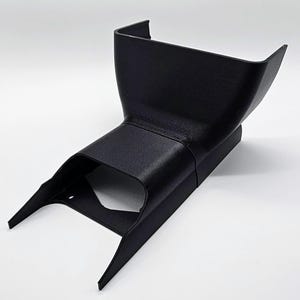 Puede incluir: Un componente de plástico negro angular con una superficie texturizada. La pieza tiene una parte superior ancha y abierta que se estrecha hacia una base más estrecha con dos soportes angulados. El diseño sugiere una pieza funcional, posiblemente automotriz.