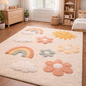 Puede incluir: Una alfombra afelpada de color crema con diseños caprichosos y coloridos. La alfombra tiene un patrón de arcoíris, soles sonrientes, nubes y flores en tonos pastel de naranja, amarillo, verde y rosa. Esta alfombra es adecuada para un dormitorio infantil.