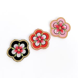 May include: Three embroidered flower patches in red, black, and pink. Each patch features a five-petal design with white and red details, outlined with gold and green stitching. The patches are on a white background.