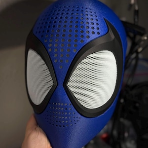 May include: A blue and black superhero mask with white mesh eye coverings. The mask has a textured surface and small circular ventilation holes on the top. The eye openings are outlined in black.