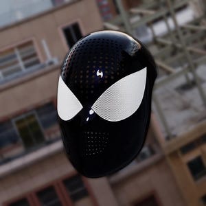 May include: A black mask with white eye coverings and a perforated design. The mask has a glossy finish and a small emblem on the forehead. The eye coverings are white with a perforated pattern. The mask is likely a costume or cosplay item.