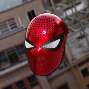 Op de afbeelding: Een glanzend rood masker met een ontwerp dat doet denken aan een superheld. Het masker heeft witte oogvormen met zwarte contouren en kleine cirkelvormige perforaties. Het masker is ontworpen om het hele gezicht te bedekken.