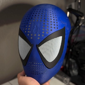 May include: A blue superhero mask with a perforated surface. The mask features black eye surrounds and white mesh eye coverings. A spider emblem is visible on the forehead. The mask is held in a hand.