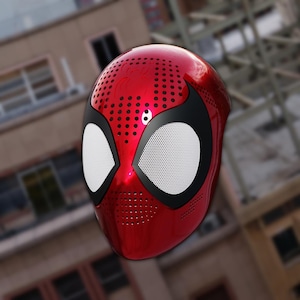 Könnte beinhalten: Eine rote Spider-Man-Maske mit schwarzen Akzenten und weißen Mesh-Augenabdeckungen. Die Maske hat eine glänzende Oberfläche und kleine Belüftungslöcher. Das Design ist vom Superhelden Spider-Man inspiriert.