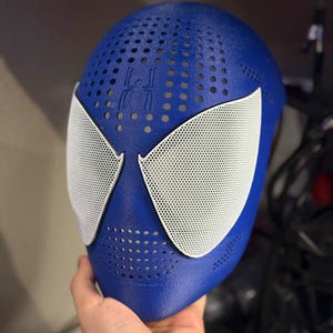 May include: A blue full-face mask featuring a textured surface with numerous small ventilation holes. The mask includes two large, white, mesh-covered eye openings and a small, embossed spider emblem centered on the forehead area above the eyes.
