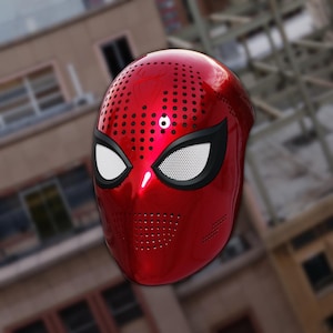 Op de afbeelding: Een glanzend rood superheldenmasker met zwarte oogomrandingen en witte mesh oogbedekkingen. Het masker heeft talrijke kleine ventilatiegaten en een glad, reflecterend oppervlak. Het ontwerp doet denken aan een populair stripboekpersonage.