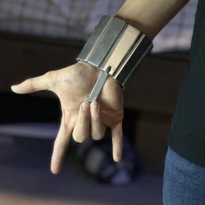May include: A silver wrist-mounted device with a rectangular design, featuring multiple parallel bars. A metal bar extends from the device, and the hand is making a rock and roll gesture. The person is wearing a black top and blue jeans.