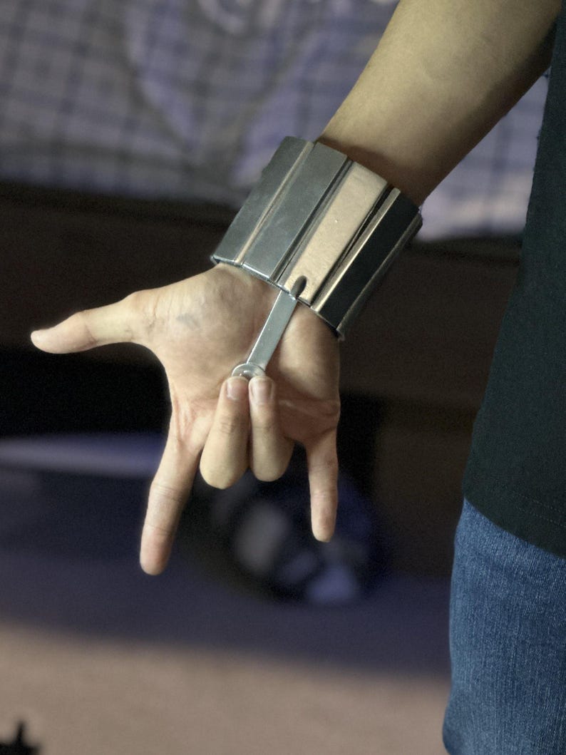 May include: A silver wrist-mounted device with a rectangular design, featuring multiple parallel bars. A metal bar extends from the device, and the hand is making a rock and roll gesture. The person is wearing a black top and blue jeans.