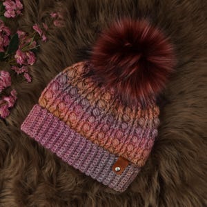 May include: A hand-knitted beanie in shades of pink, orange, and purple, topped with a large, fluffy burgundy pom-pom. The hat features a textured stitch pattern and a small brown leather tag. The beanie is displayed on a brown, textured surface.