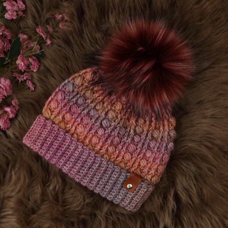 May include: A hand-knitted beanie in shades of pink, orange, and purple, topped with a large, fluffy burgundy pom-pom. The hat features a textured stitch pattern and a small brown leather tag. The beanie is displayed on a brown, textured surface.