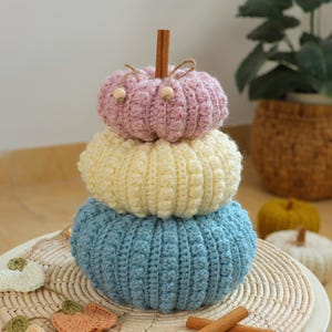 May include: A stacked trio of crocheted pumpkins in pastel pink, cream, and light blue, topped with a cinnamon stick stem and small wooden beads on twine. The pumpkins feature a textured bobble stitch pattern and are arranged vertically.