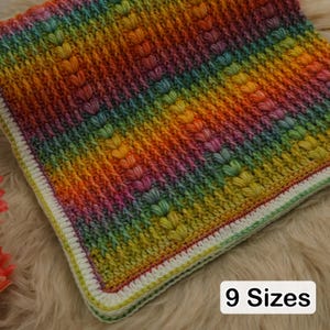May include: A colourful, square blanket in a rainbow gradient. The blanket has a textured, raised pattern and a white and yellow border. The text "9 Sizes" is displayed in a white box in the lower right corner.
