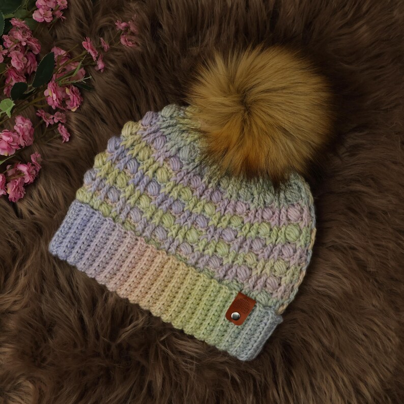 May include: A hand-knitted beanie hat with a gradient of pastel colors, including lavender, yellow, and peach. The hat features a textured stitch pattern and a large, fluffy, brown faux fur pom-pom on top. A small, brown leather tag is sewn on the brim.