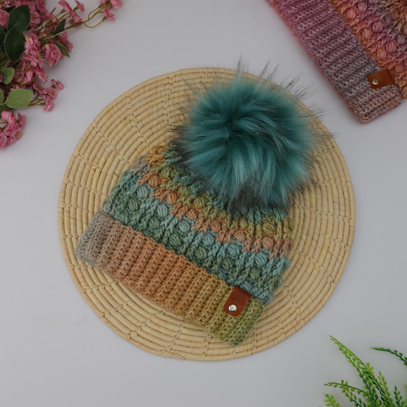 May include: A handmade, multicolored knit hat with a teal faux fur pom-pom. The hat features a textured pattern in shades of brown, green, and blue. A matching knit cuff is also visible. A small brown leather tag is attached to the hat.