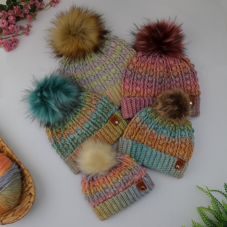 May include: A collection of colorful, hand-knitted beanies with faux fur pom-poms. The hats feature a textured stitch pattern and are in various pastel shades, including pink, blue, yellow, and green. Each hat has a small, brown leather tag.