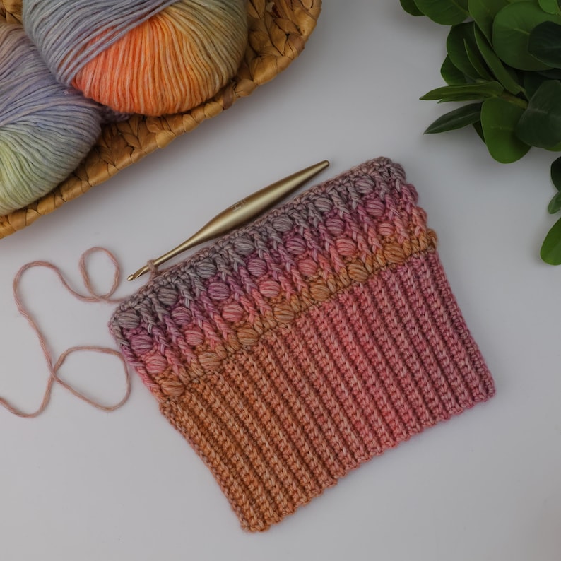 May include: A handmade crochet project in progress, featuring a hat with a textured, bobble stitch design. The hat transitions from orange to pink and purple hues. A gold crochet hook and colorful yarn balls in a wicker basket are also visible.