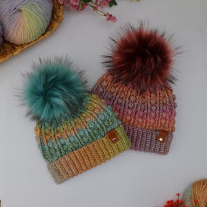May include: Two handmade knit beanies with large faux fur pom-poms. One beanie is green, yellow, and blue, the other is pink, orange, and purple. Both have a textured stitch pattern and a small leather tag.
