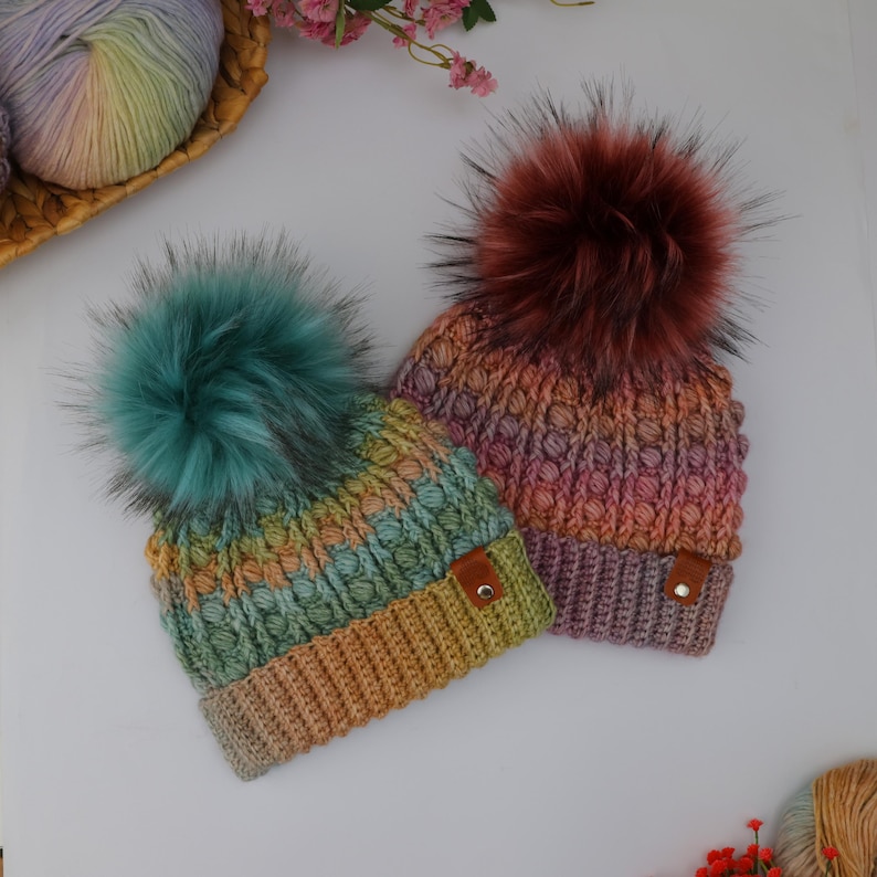 May include: Two handmade knit beanies with large faux fur pom-poms. One beanie is green, yellow, and blue, the other is pink, orange, and purple. Both have a textured stitch pattern and a small leather tag.