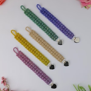 May include: An overhead shot displays six handmade crochet pacifier clips in various colors: purple, yellow, beige, green, and blue. Each clip features a textured stitch pattern, a loop at one end, and a heart-shaped metal clip at the other.