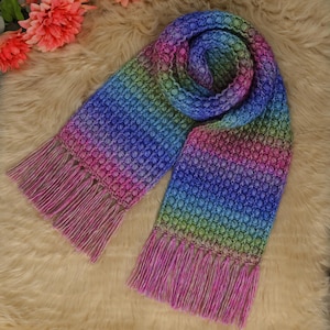 May include: A multi-coloured knitted scarf with a textured pattern and fringed ends. The scarf features a gradient of colours, including pink, blue, green, and purple. It is displayed on a soft, light-coloured surface.