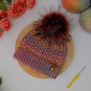 May include: A handmade, multi-colored knit hat with a large, fluffy, burgundy faux fur pom-pom. The hat features a textured stitch pattern and a brown leather tag. A yellow crochet hook rests nearby.