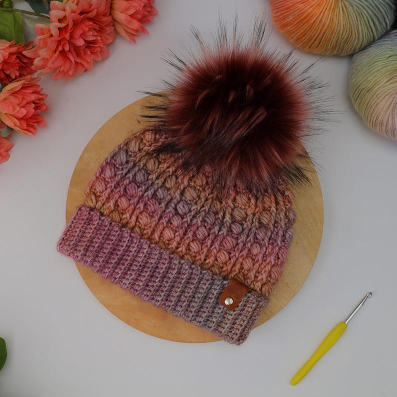 May include: A handmade, multi-colored knit hat with a large, fluffy, burgundy faux fur pom-pom. The hat features a textured stitch pattern and a brown leather tag. A yellow crochet hook rests nearby.