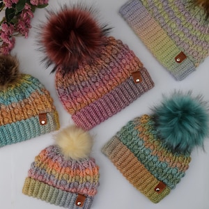 Op de afbeelding: Vijf kleurrijke gebreide mutsen met imitatiebont pompons. Elke muts heeft een unieke kleurovergang, variërend van blauw en groen tot oranje en paars. De mutsen hebben een gestructureerd steekpatroon en een klein leren label.