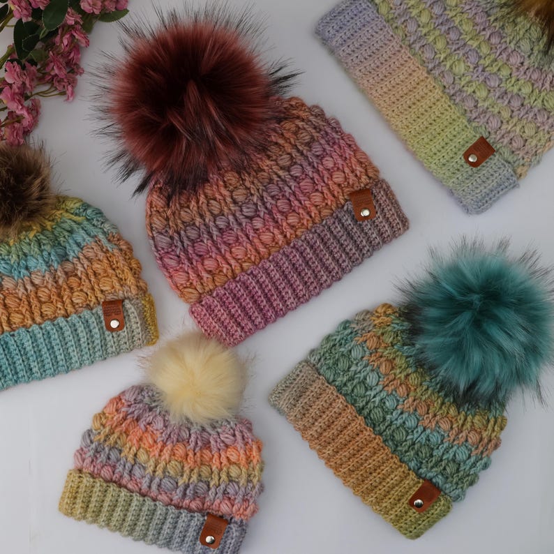 May include: Five colorful knit beanies with faux fur pom-poms. Each hat features a unique color gradient, ranging from blues and greens to oranges and purples. The hats have a textured stitch pattern and a small leather tag.