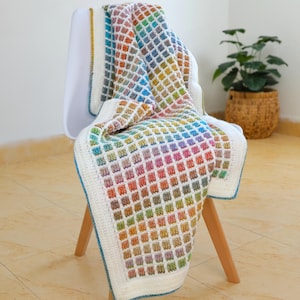 May include: A colorful, handmade crochet blanket draped over a white chair. The blanket features a repeating square pattern in a rainbow of colors, with a white border and blue trim. The blanket is a cozy home decor item.