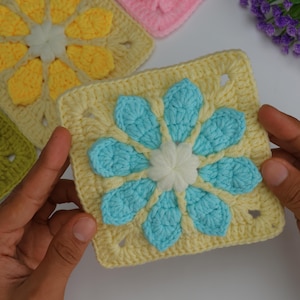May include: A close-up of a crocheted square with a flower design. The square is light yellow with a blue flower and a white center. Other squares with different colored flowers are in the background.