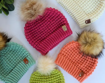 May include: Five crocheted beanies in cream, magenta, mint green, lime green, and coral. Each hat features a textured bobble stitch pattern, a wide ribbed cuff, a faux fur pom-pom on top, and a small brown leather-like tag with a metal rivet.