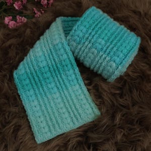 May include: A turquoise and aqua knitted scarf and headband set. The scarf and headband have a textured pattern with vertical ribbing and a leaf-like design. The set is displayed on a brown, textured surface, creating a cozy aesthetic.