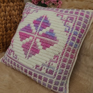以下が含まれることがあります： ピンクと紫の色合いの幾何学模様が施された、正方形のオフホワイトのかぎ針編み枕。中央のモチーフは4枚の花びらの花の形です。ボーダーは正方形の繰り返し模様です。枕は明るい茶色の表面にあります。