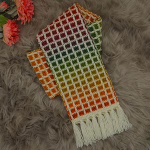 Op de afbeelding: Een handgemaakte, kleurrijke sjaal met een wafelachtig patroon. De sjaal gaat over in een regenboog van kleuren, van oranje tot rood tot groen, en heeft witte franjes aan de uiteinden. De sjaal is gemaakt van zacht garen.