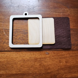 May include: A wooden square frame with a smaller square cutout in the center. The frame is on top of a smaller square piece of wood with a brown felt backing.