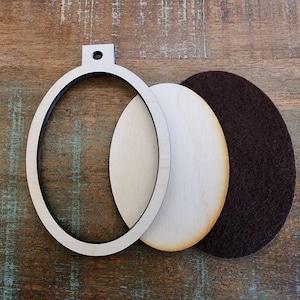 May include: Oval-shaped wooden craft supplies, including a frame, two wooden inserts, and a brown felt backing. The frame has a small hole at the top for hanging. The items are arranged on a rustic wooden surface.