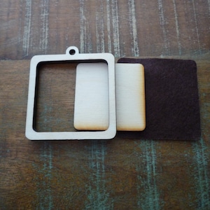 May include: A white wooden frame with a cutout for a picture or embroidery. The frame is attached to a white backing board and a brown felt backing.