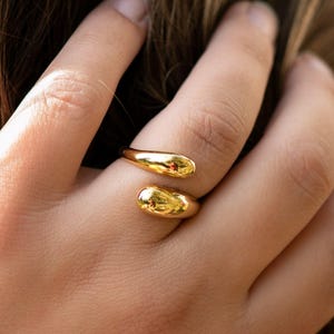 Könnte beinhalten: Ein goldfarbener Ring mit offenem Design, der zwei abgerundete Enden aufweist, die den Finger teilweise umschließen. Ein Ende hat einen kleinen, roten Akzent. Der Ring wird an einer Hand mit langen, hellbraunen Haaren im Hintergrund gezeigt.