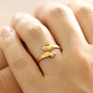 Könnte beinhalten: Ein goldfarbener Ring mit einem Design aus zwei sich umarmenden Händen. Der Ring wird an einem Finger getragen, wobei die Hände scheinbar den Finger umschließen. Das Design des Rings ist detailliert und scheint aus Metall zu sein.