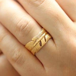 Peut inclure: Une bague dorée avec un motif de plume. La bague, portée au doigt, présente des motifs de plumes détaillés. La conception comprend une veine centrale et des détails latéraux, créant un aspect de plume réaliste.