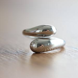 Peut inclure: Gros plan d'une bague argentée au design unique. La bague présente deux éléments arrondis en forme de goutte d'eau qui se chevauchent. Le métal poli reflète l'environnement, créant un aspect brillant. La bague est posée sur une surface en bois.