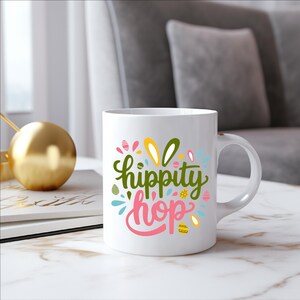 A white coffee mug with a colorful design featuring the text "hippity hop" on it, placed on a marble surface next to a gold ornament.