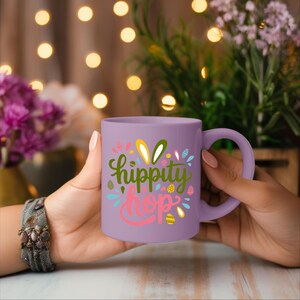 A person's hand holding a purple mug with the text "hippity hop" printed on it, with a background of colorful lights and flowers.
