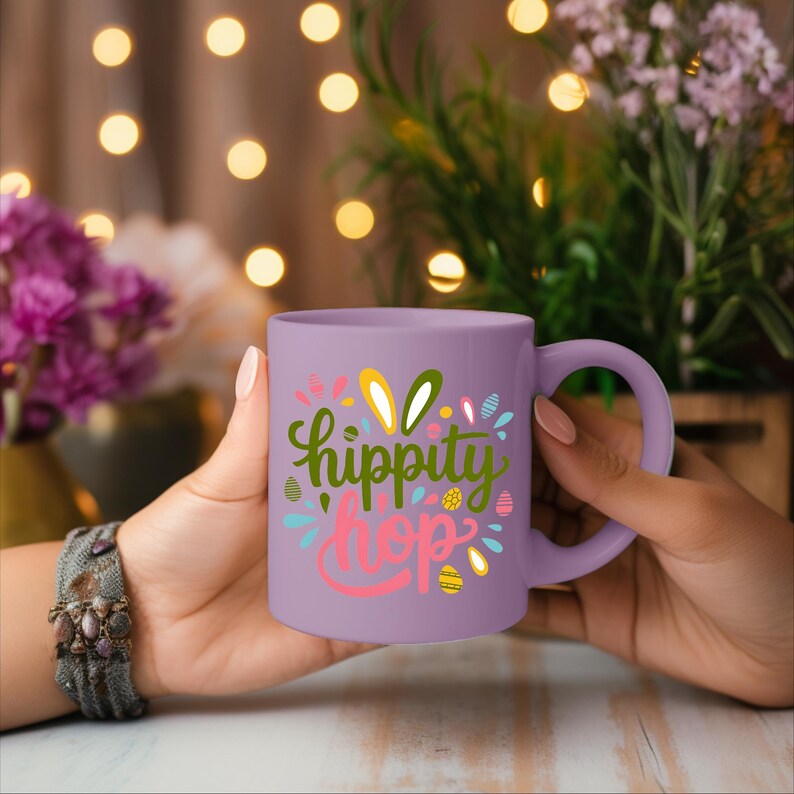 A person's hand holding a purple mug with the text "hippity hop" printed on it, with a background of colorful lights and flowers.